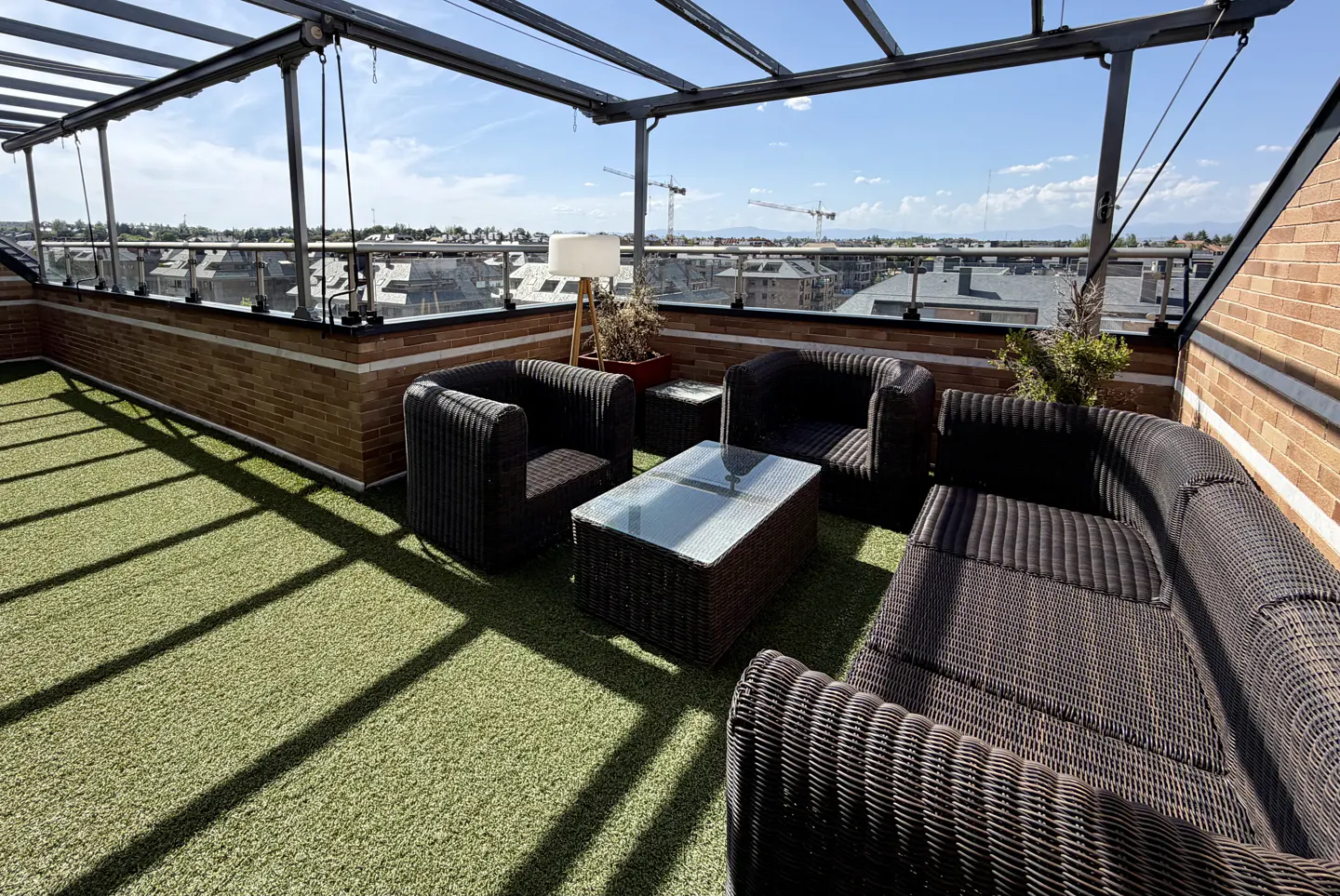 Rooftop patio with wicker furniture, glass table, and artificial grass. Cityscape visible in the background under a blue sky.