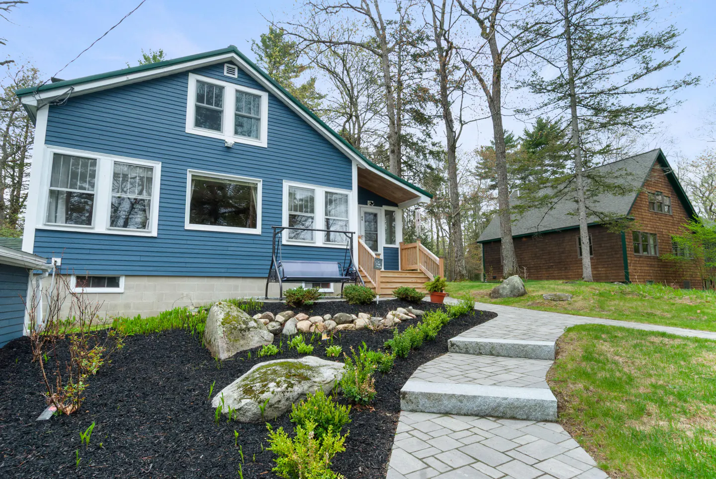 Blue house with white trim and green roof, stone walkway, and black mulch landscaping. A swing hangs on the porch.