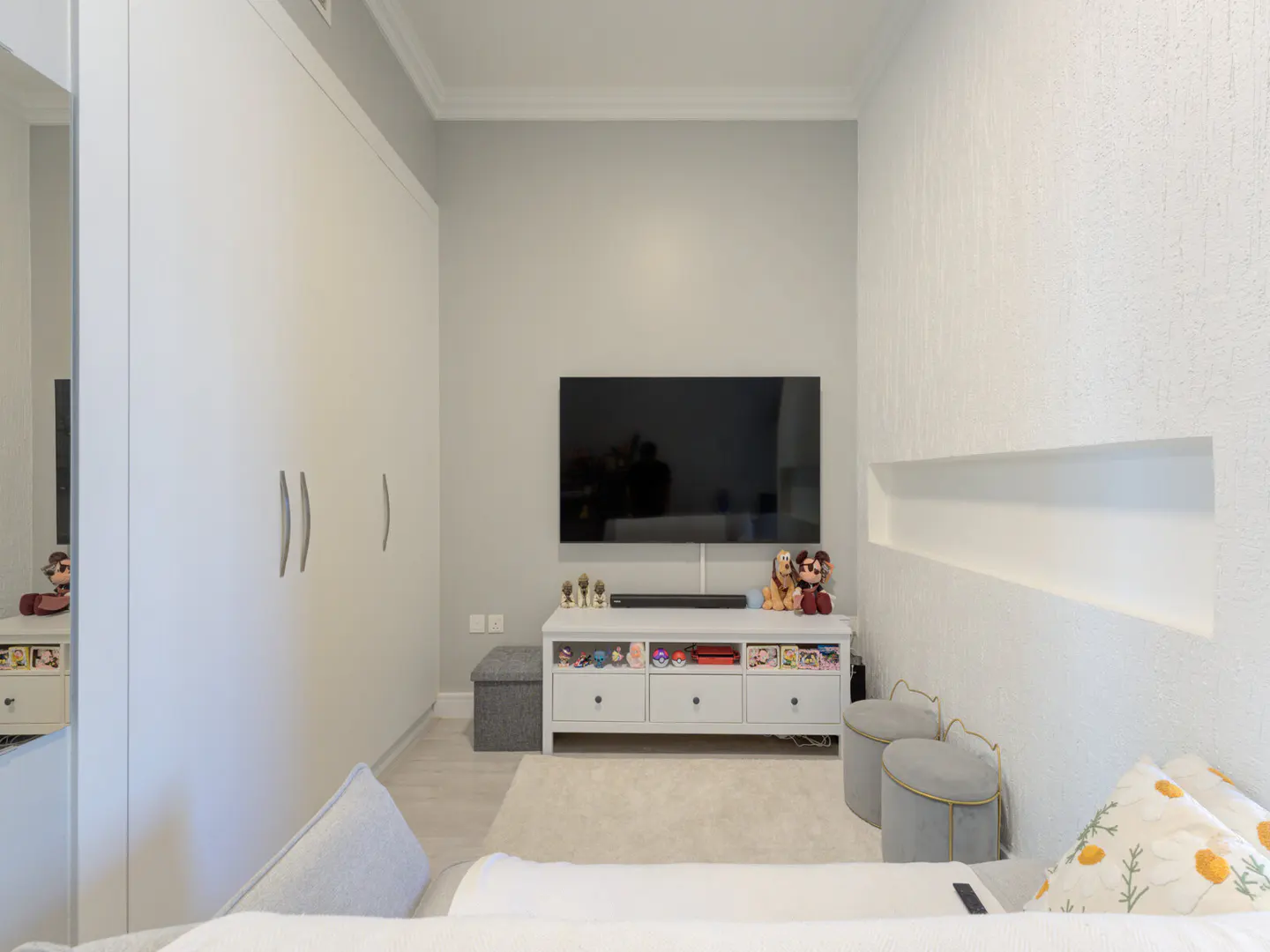 A bedroom with a TV, white cabinets, a white console table, and two gray stools.
