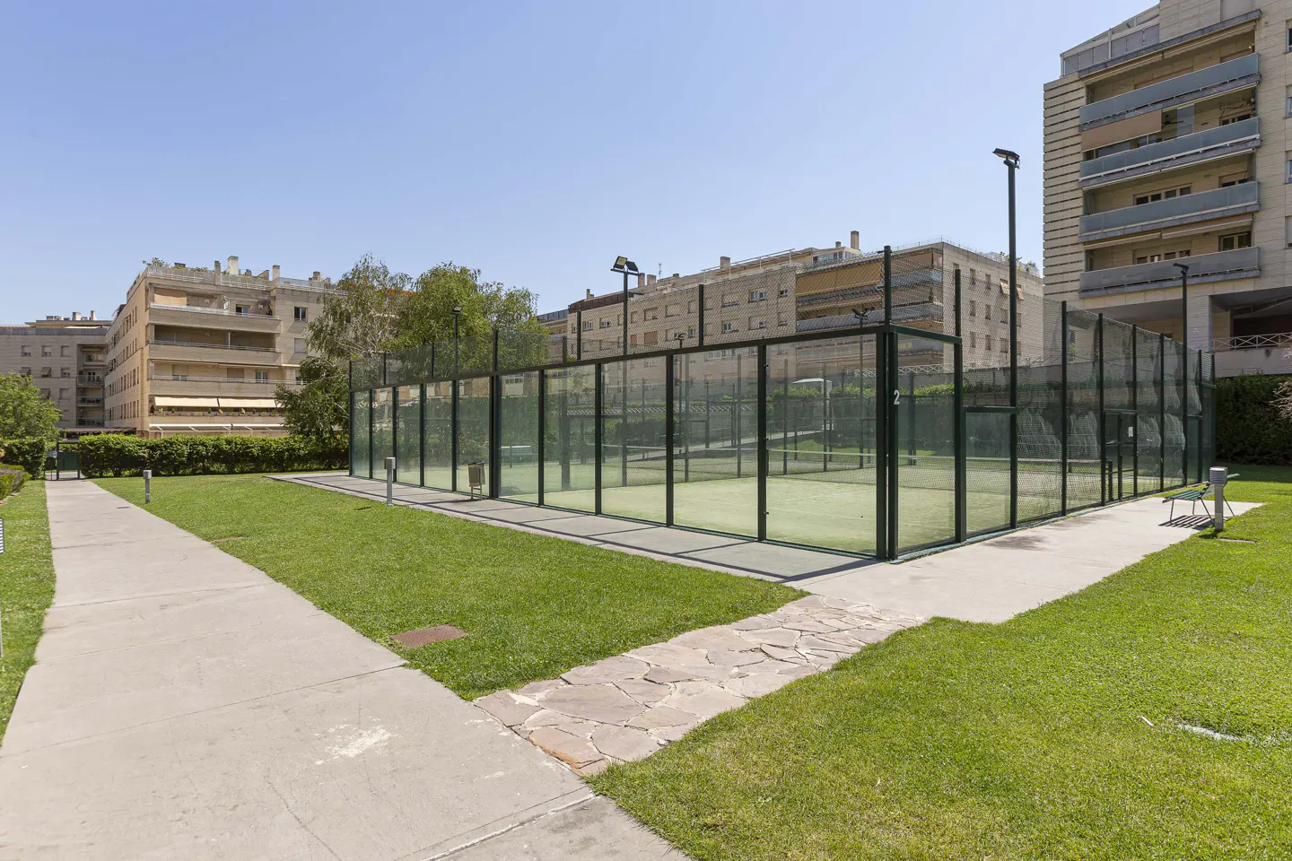Outdoor paddle tennis court with glass walls, green turf, and a concrete walkway, set against a backdrop of apartment buildings under a blue sky.