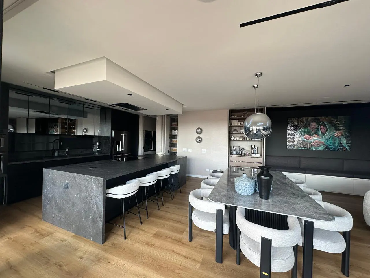A modern kitchen and dining area with a long, gray island and white stools. A dining table with white chairs sits under a chrome pendant light.