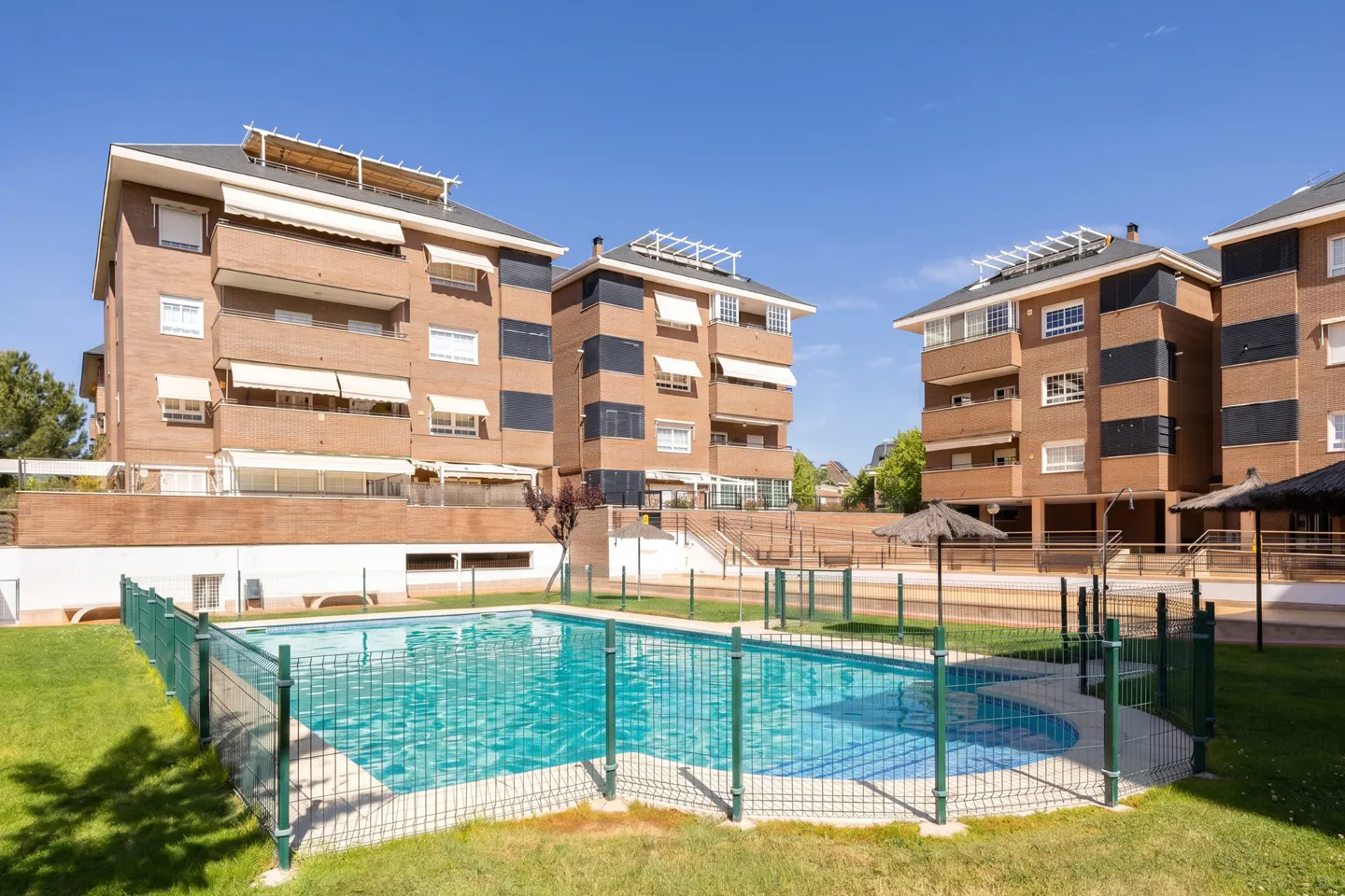 Exterior view of brick apartment buildings with a turquoise swimming pool and green fence on a sunny day.