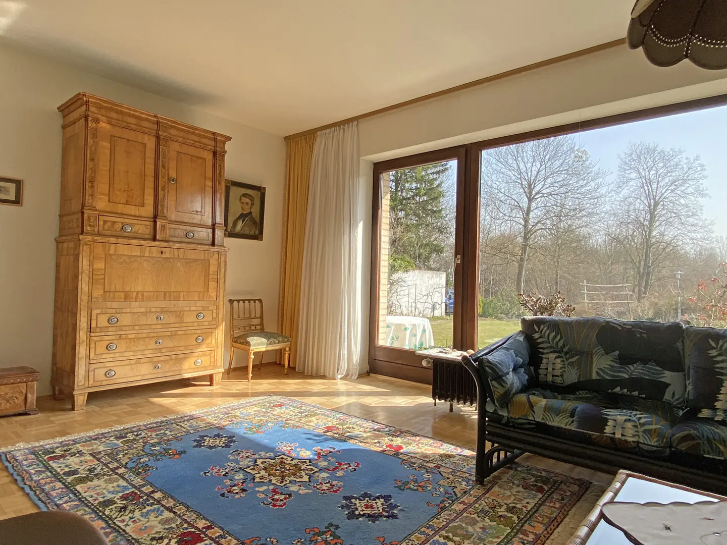 Bright living room with a large window, blue floral rug, wooden cabinet, and a floral sofa. Outside is a garden with trees.