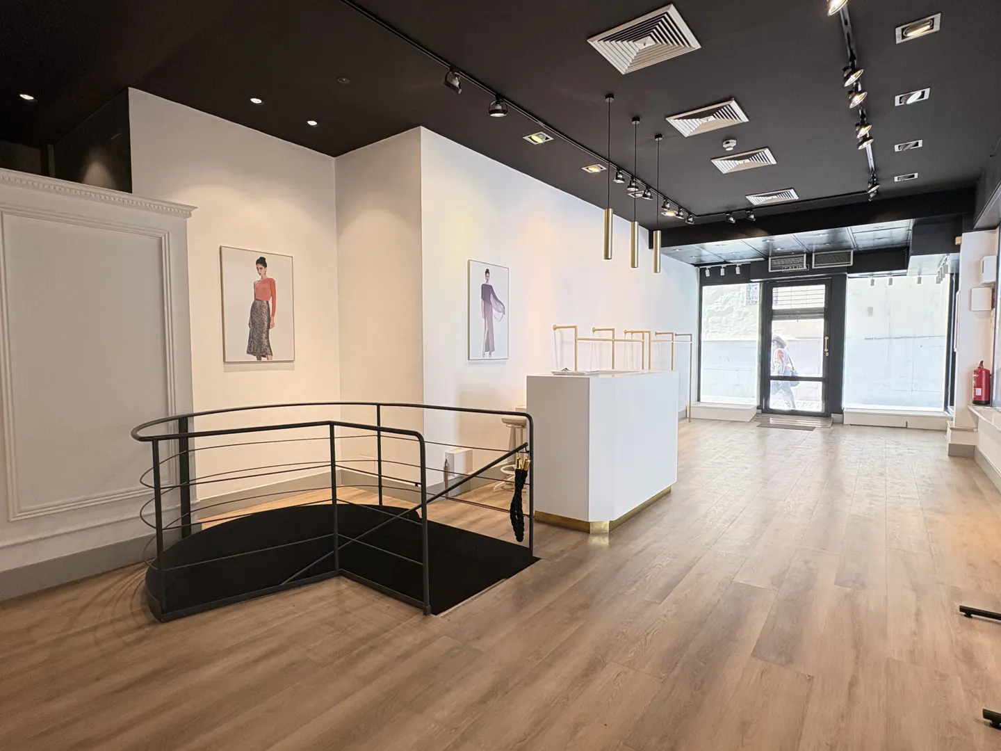 Interior of a retail space with light wood floors, white walls, and a black ceiling with track lighting. Clothing racks and framed art are visible.