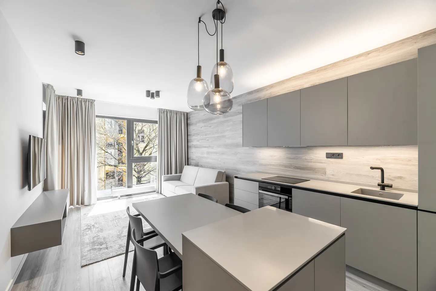 Modern apartment interior with gray cabinets, island, dining table, and sofa. Large window with curtains lets in natural light.