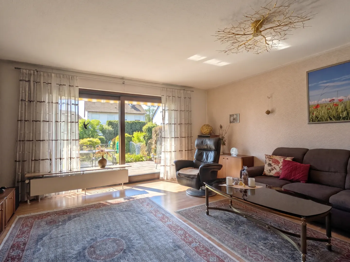 Bright living room with brown sofa, leather recliner, and glass coffee table. Sliding glass doors open to a green garden.