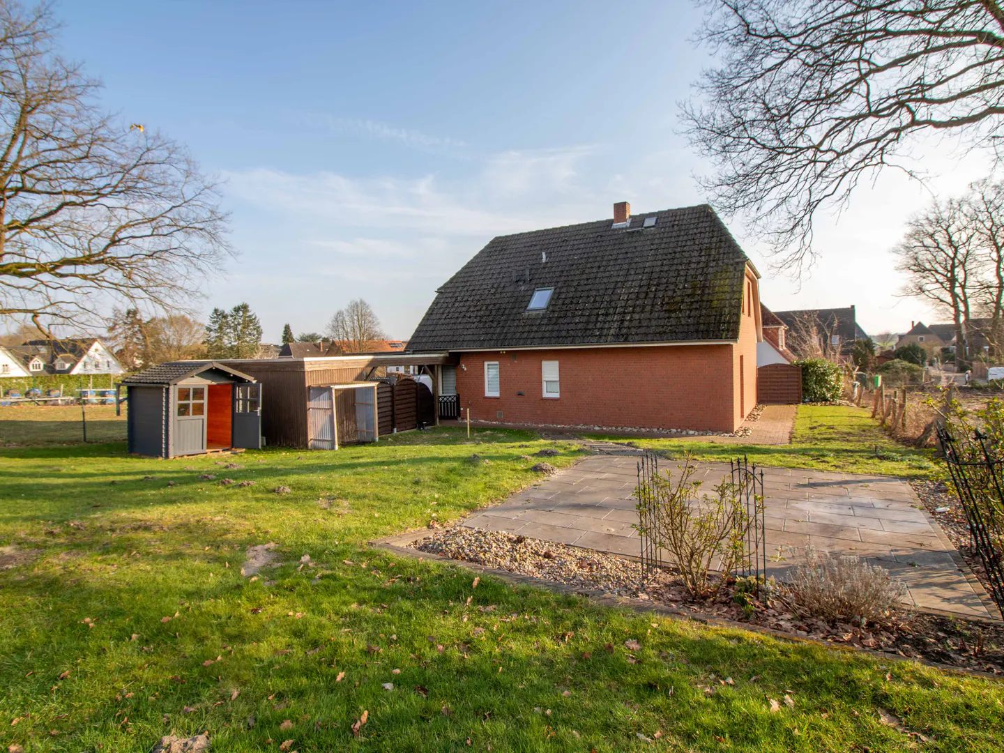 Exterior view of a red brick house with a dark roof, green lawn, and storage sheds on a sunny day.