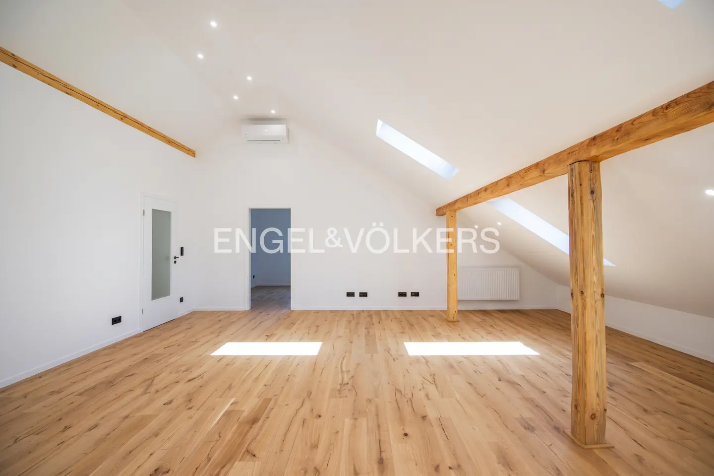 Bright attic room with wood floors, white walls, and exposed wooden beams. Skylights provide natural light. Engel & Völkers logo visible.