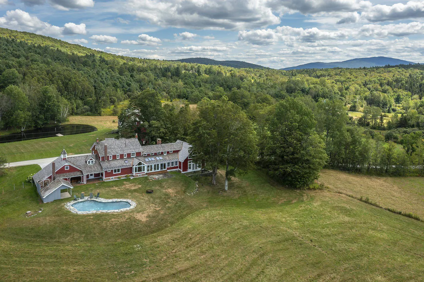 Aerial view of a red farmhouse with a pool, surrounded by green fields and trees, with mountains in the background.