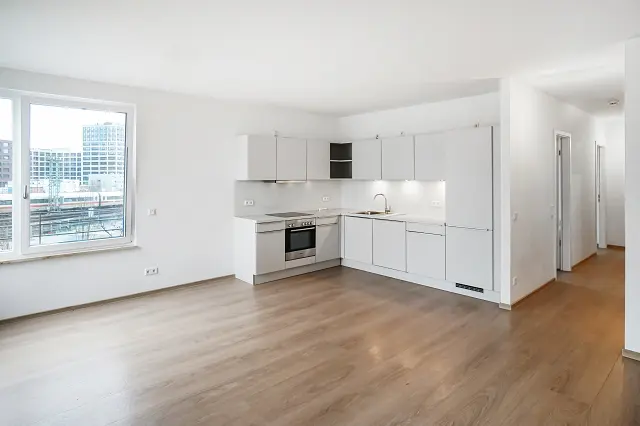 Bright, modern apartment with wood floors and white walls. A white kitchen features stainless steel appliances. A large window overlooks a city view.
