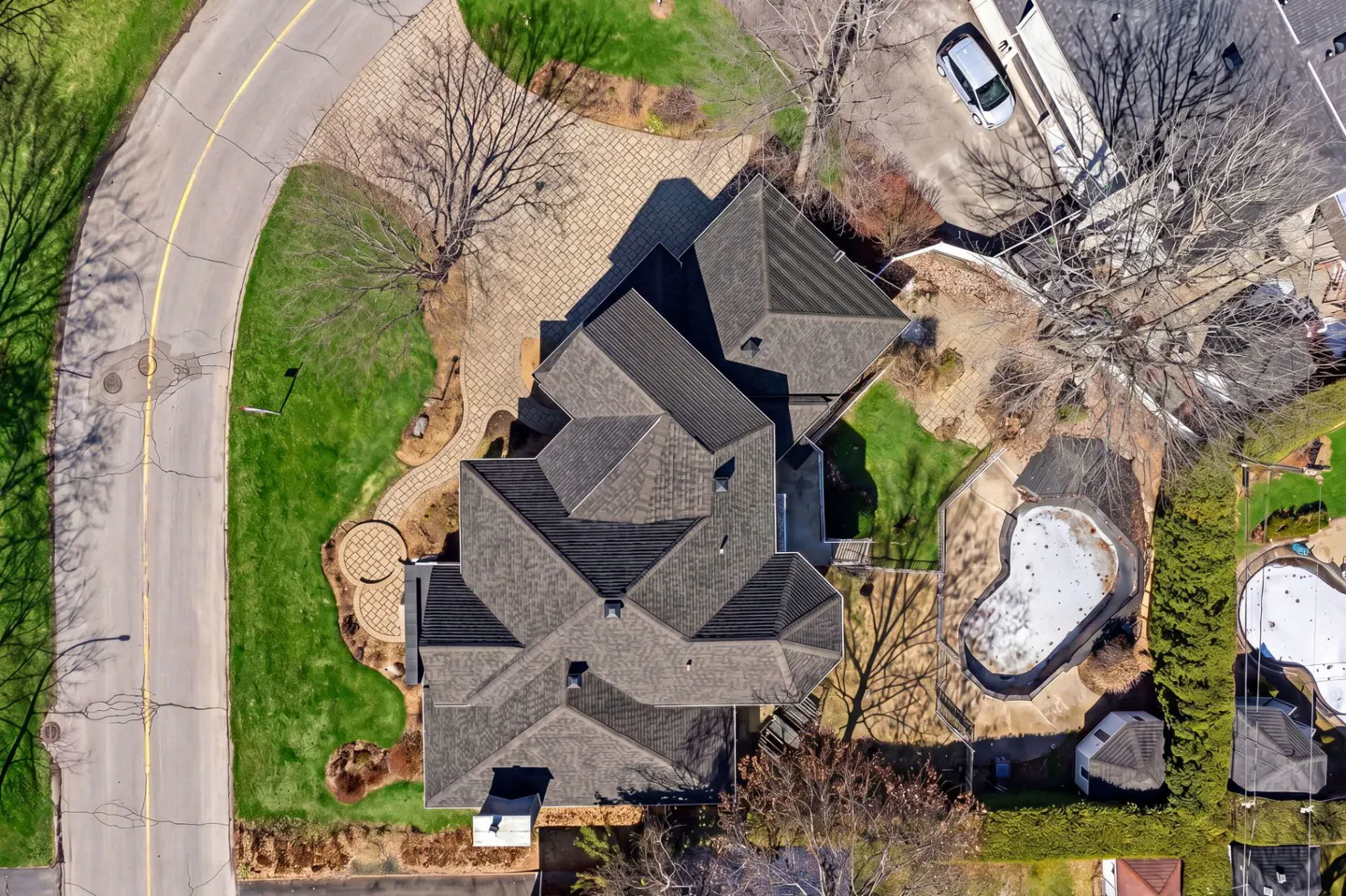 Aerial view of a large house with a complex gray roof, green lawn, and a kidney-shaped pool covered for the winter.