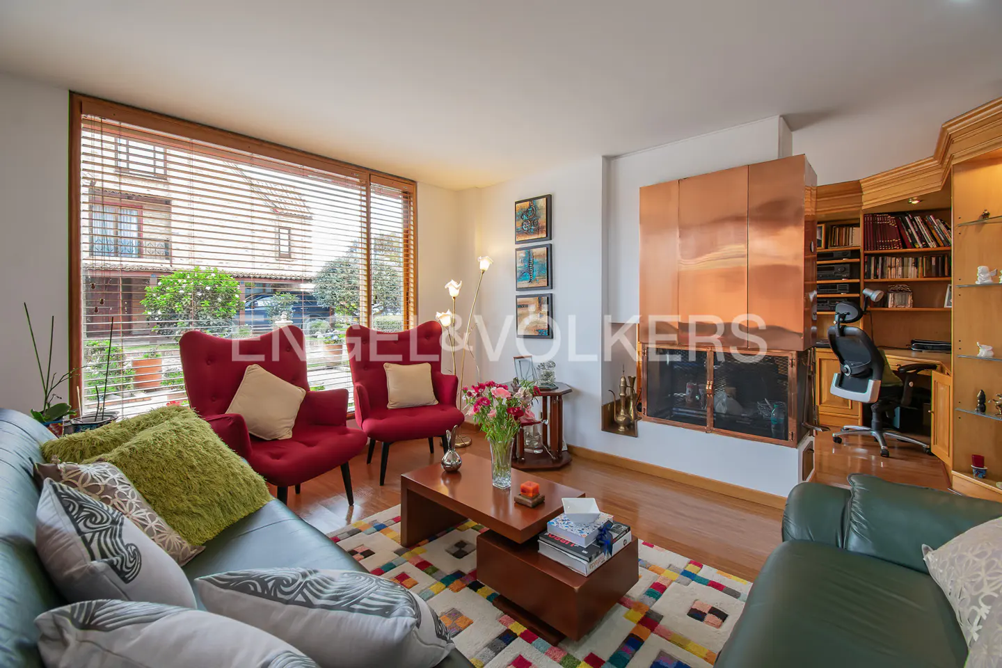 Living room with red chairs, green sofas, wood floors, and a copper fireplace. A colorful rug sits in the center.