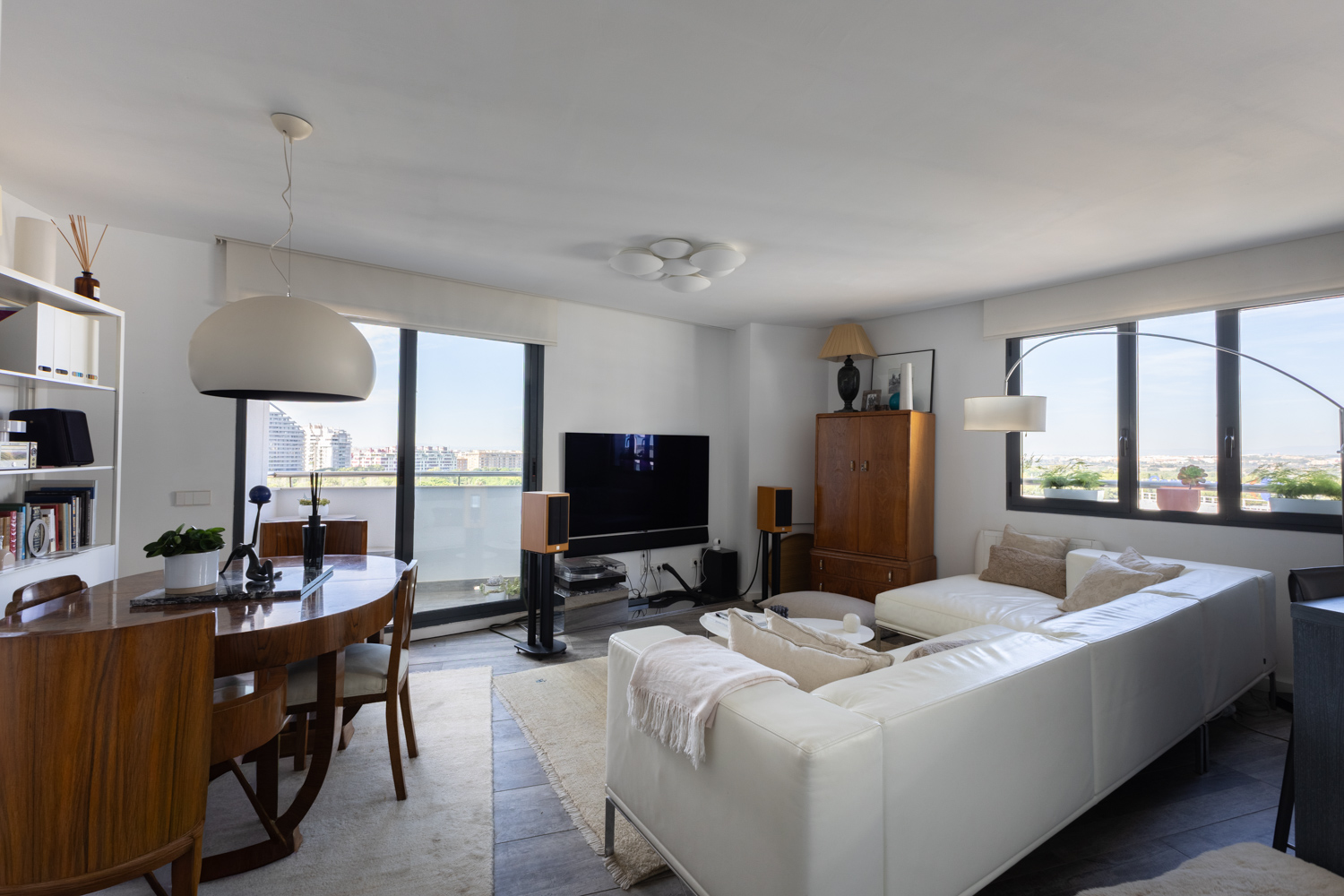 Bright living room with a white sectional sofa, wooden dining set, large windows, and a wall-mounted TV. [AI-generated text]