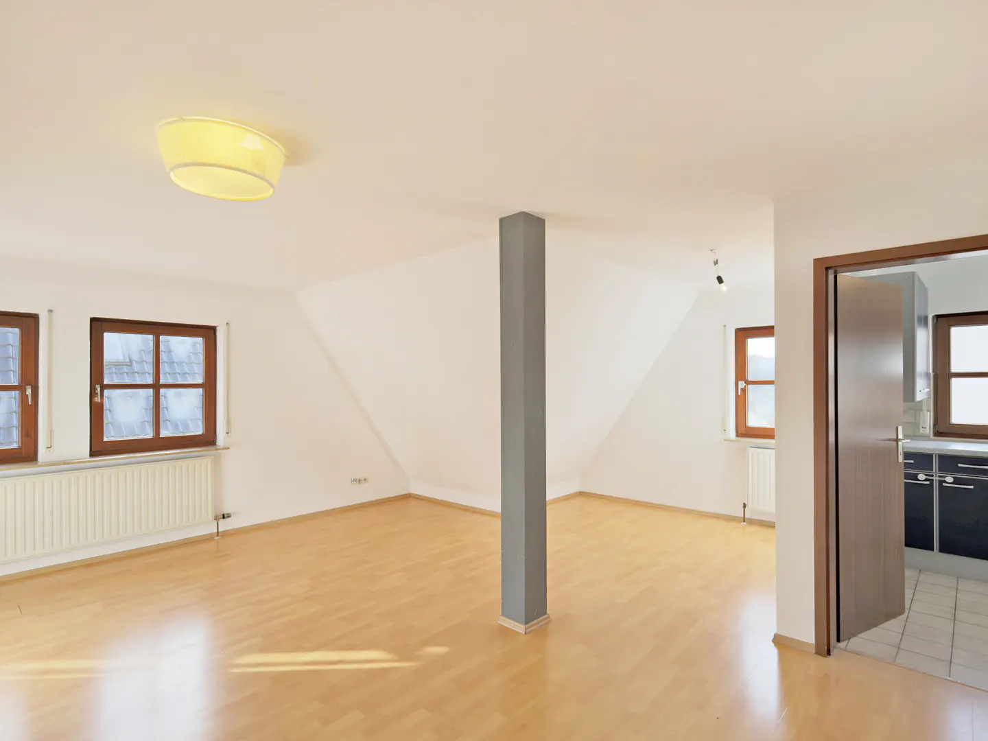 Bright, empty room with wood floors, white walls, and a gray support column. Windows with wood frames and a doorway to a kitchen are visible.