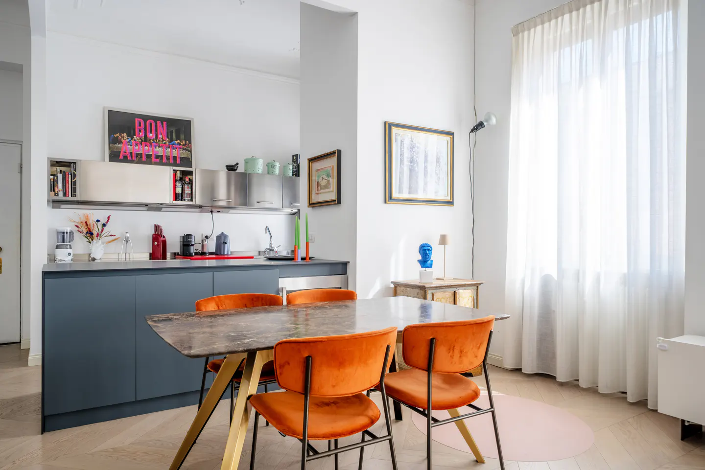 A modern kitchen and dining area with blue cabinets, orange chairs, and a marble table. White walls and sheer curtains create a bright, airy space.