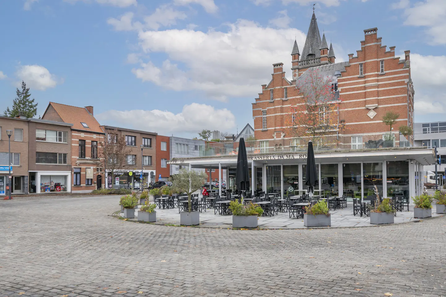A cobblestone town square featuring a modern restaurant terrace with outdoor seating in front of a historic brick building.