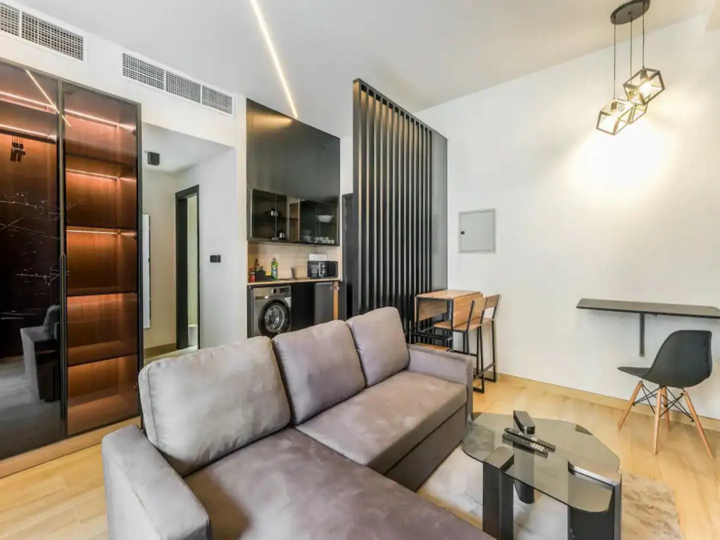 A modern studio apartment with a gray sofa, glass coffee table, and a small kitchen area.