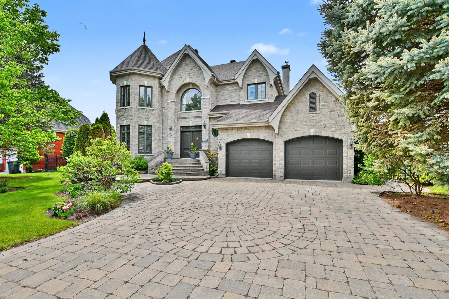 Two-story stone house with a turret, double garage, and circular paved driveway surrounded by lush green landscaping.
