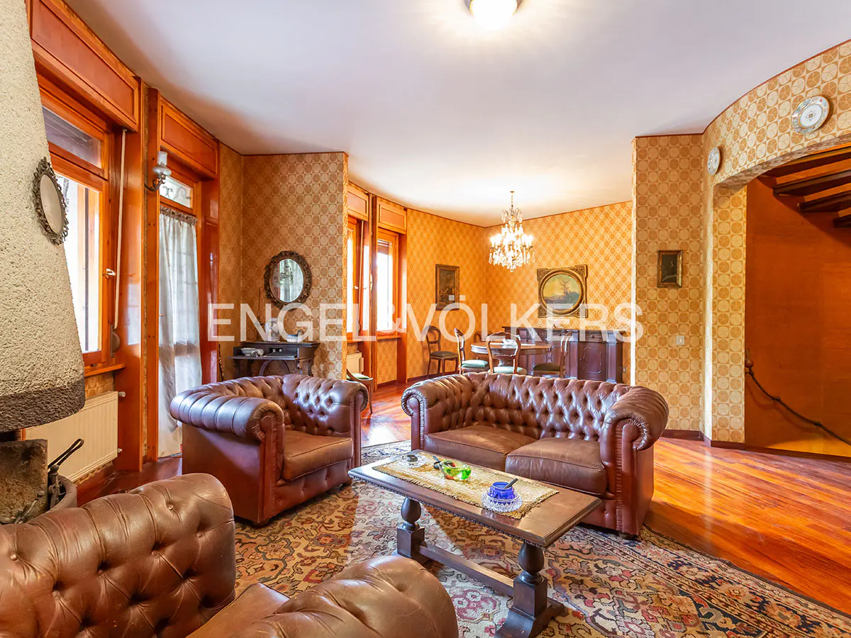 A living room with brown leather sofas, a wooden table, and patterned wallpaper. A chandelier hangs from the ceiling.