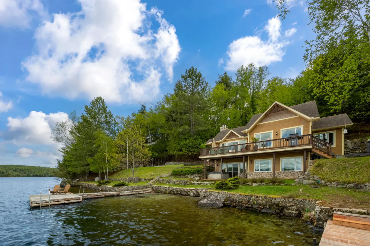 Tan lakefront home with a wooden deck, stone foundation, and private dock on a sunny day with lush green trees.