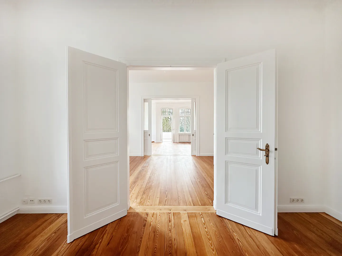 Interior view through open white double doors, revealing hardwood floors and a bright, empty room with windows.