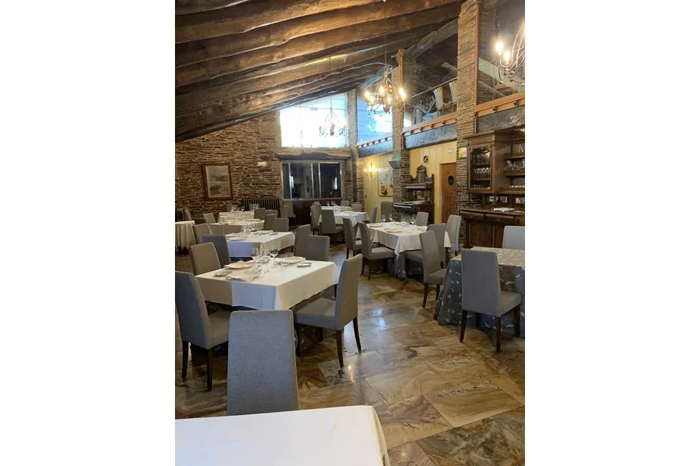 Rustic restaurant interior with stone walls, exposed wooden beams, tiled floors, and tables set with white tablecloths.
