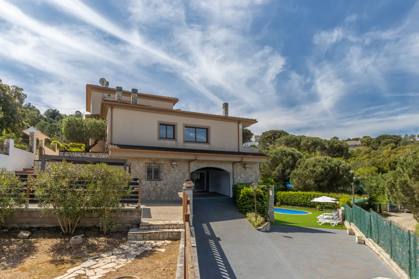 Two-story beige house with a stone facade, a driveway leading to a garage, and a pool in the backyard.