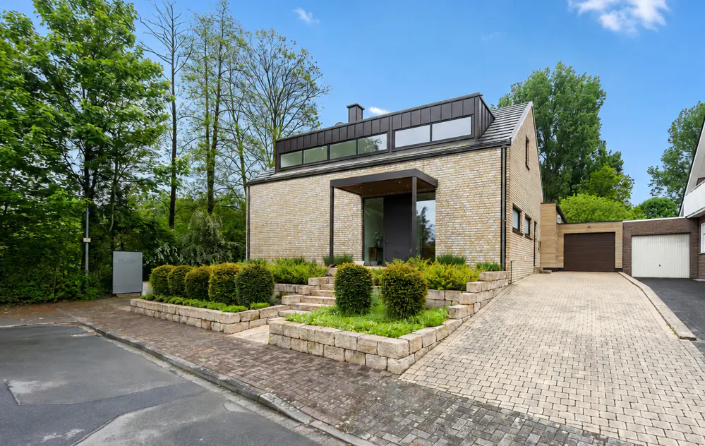 Modern beige brick house with a black roof and trim, surrounded by green trees and landscaping under a blue sky.