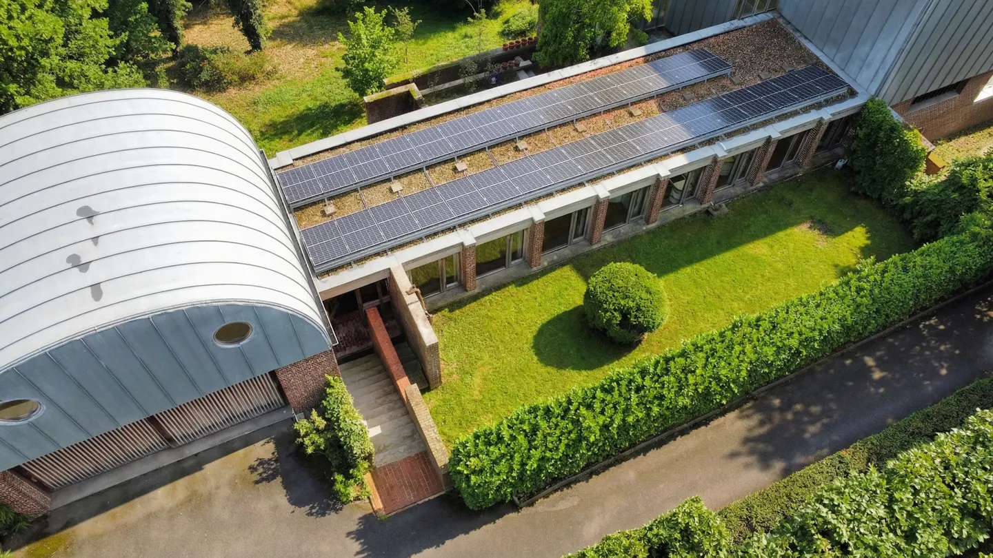 Aerial view of a modern brick building with a curved metal roof and solar panels on the flat roof, surrounded by green lawn and trees.