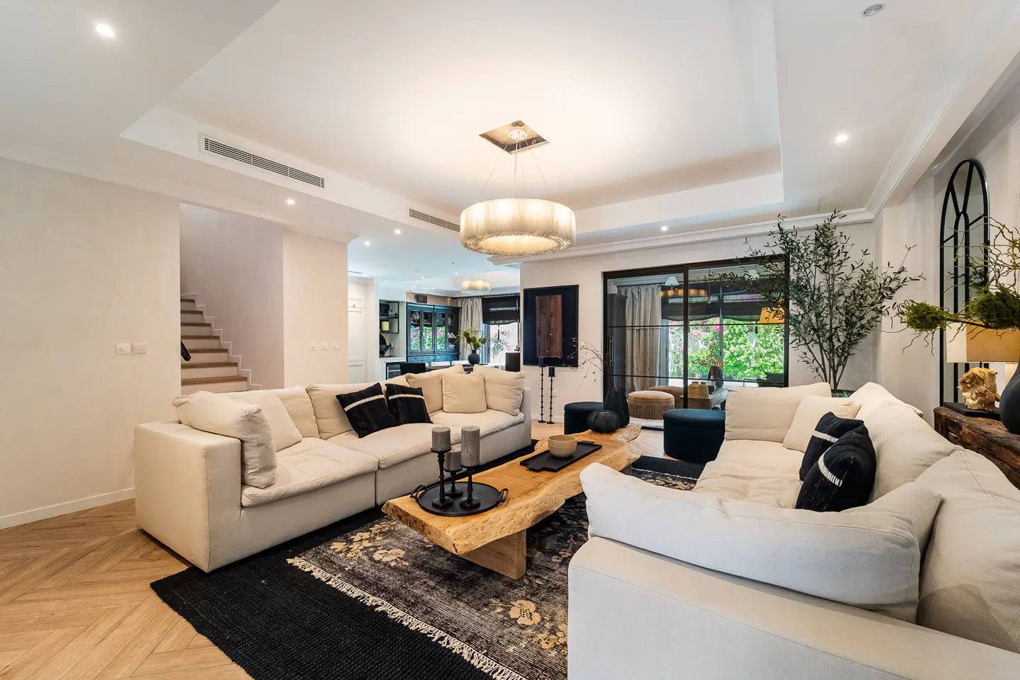 Bright living room with white sofas, black pillows, and a live-edge coffee table on a patterned rug. A staircase is visible in the background.
