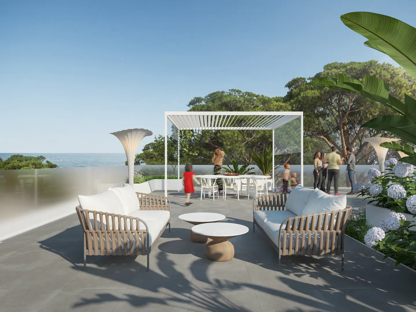 Outdoor patio with white sofas, tables, and pergola. People are gathered, with ocean and trees in the background.