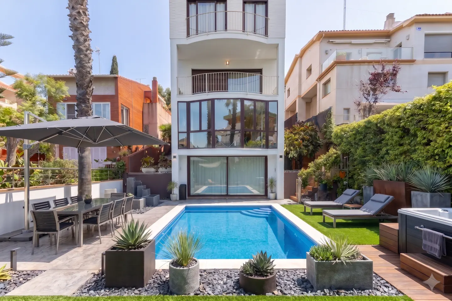 A modern white house with balconies overlooks a blue swimming pool, patio furniture, and lush greenery.