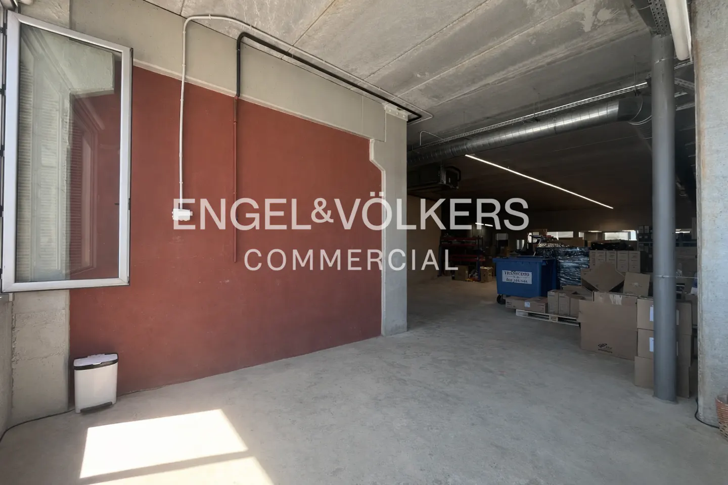 Commercial space with concrete floors and ceiling, a red wall with the Engel & Völkers logo, and boxes in the background.