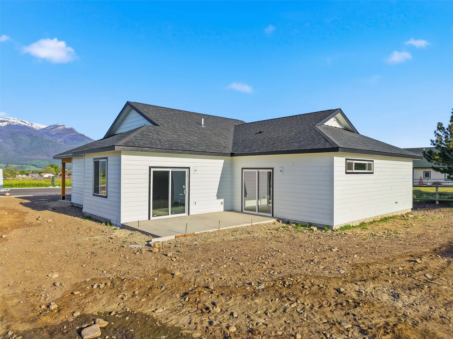 Rear view of a white, single-story house with a dark gray roof and a concrete patio. Mountains are visible in the background under a blue sky.
