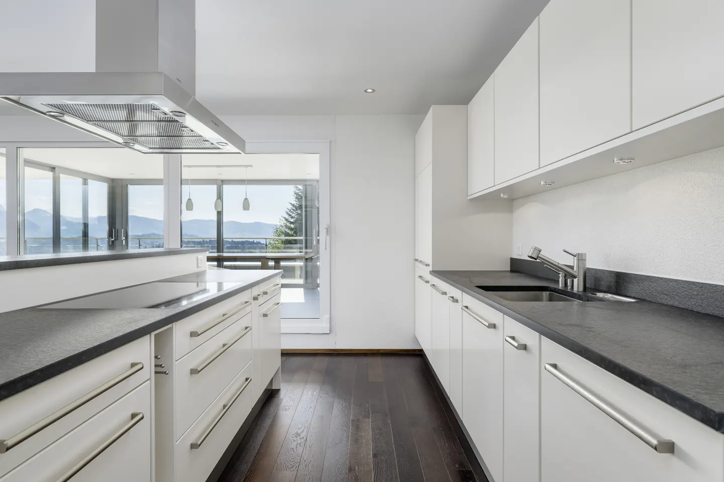 Bright kitchen with white cabinets, dark countertops, and wood floors. A stainless steel range hood hangs over the island. Large windows offer a mountain view.