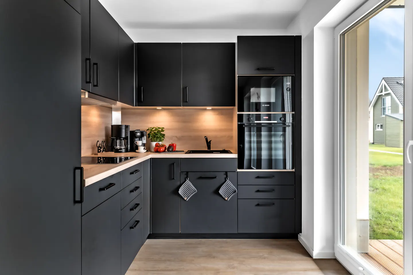 Modern kitchen with black cabinets, light wood countertops, and stainless steel appliances. A sliding glass door leads to a green lawn.