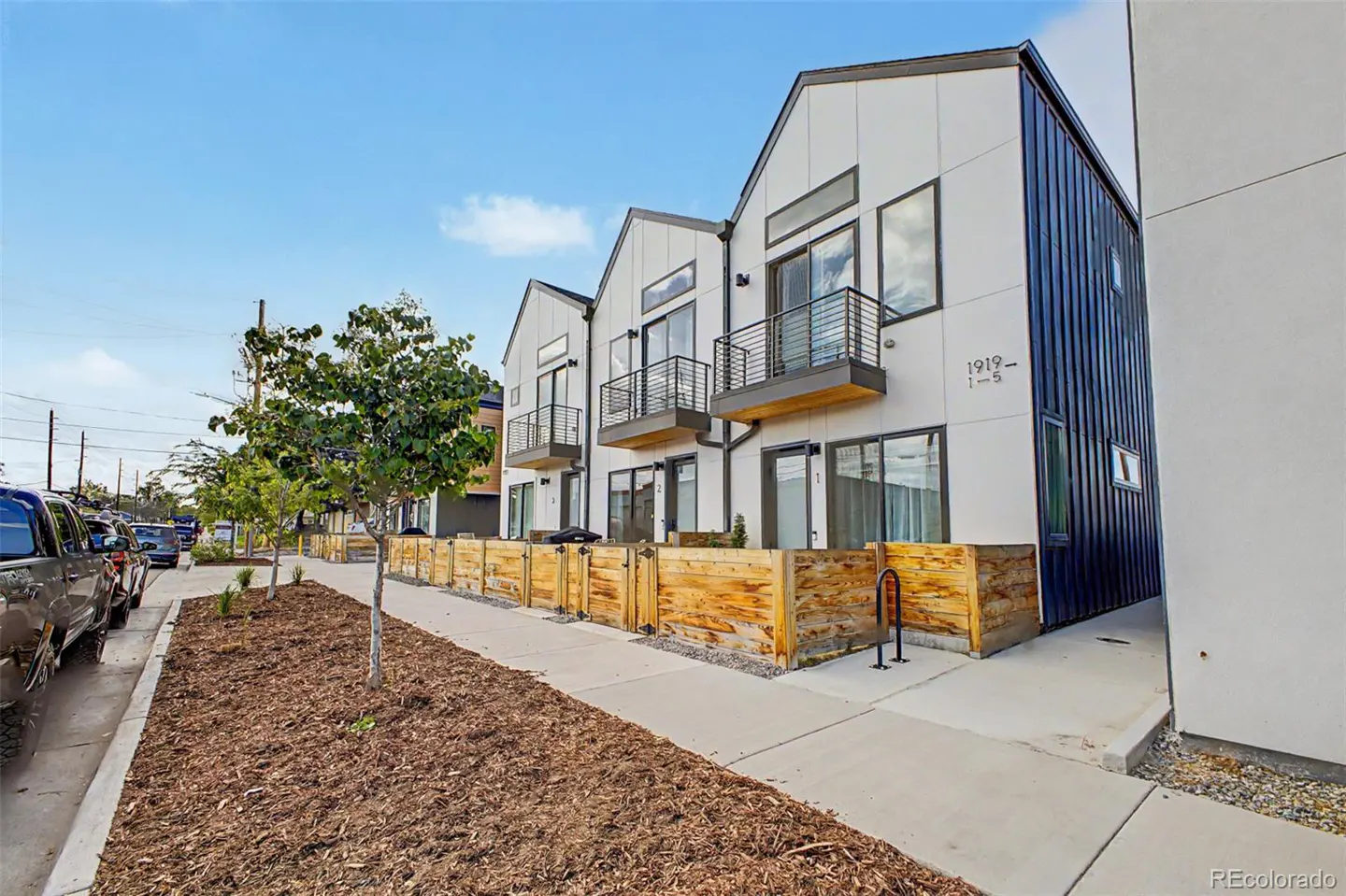 Modern townhouses with white siding, black trim, and small balconies line a sidewalk with wood fencing and young trees.