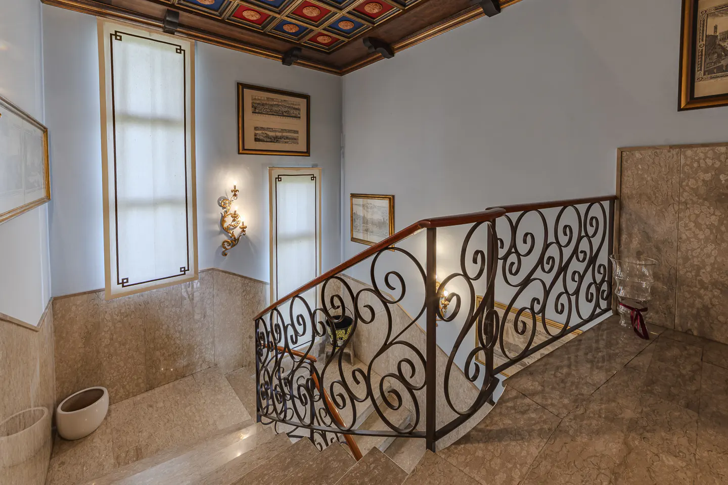 Staircase with ornate iron railing, marble floors, light blue walls, and a decorative coffered ceiling with framed art.