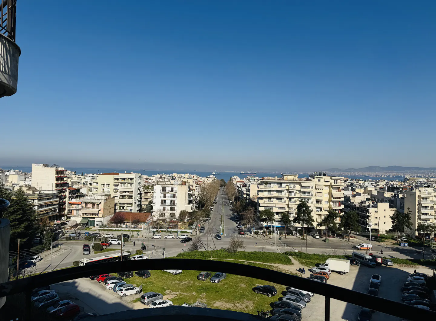 Cityscape view from a balcony, featuring buildings, a road, parked cars, and a blue sky.