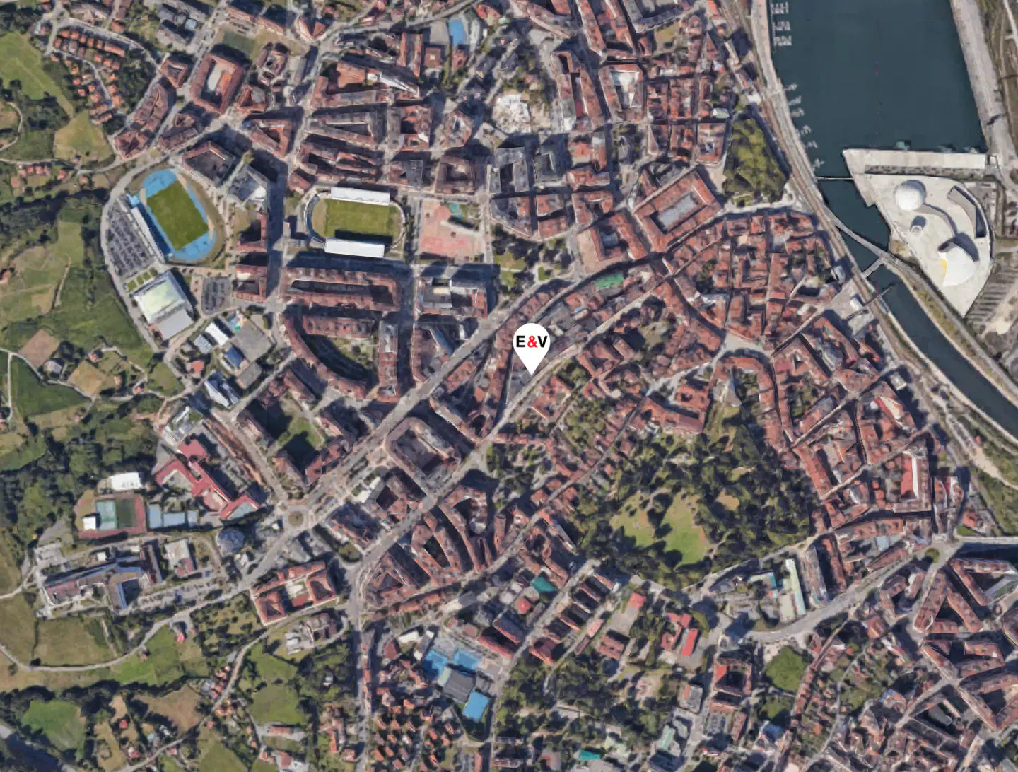 Aerial view of a city with red-roofed buildings, green spaces, a river, and a harbor. A white pin with "E&V" marks a location.