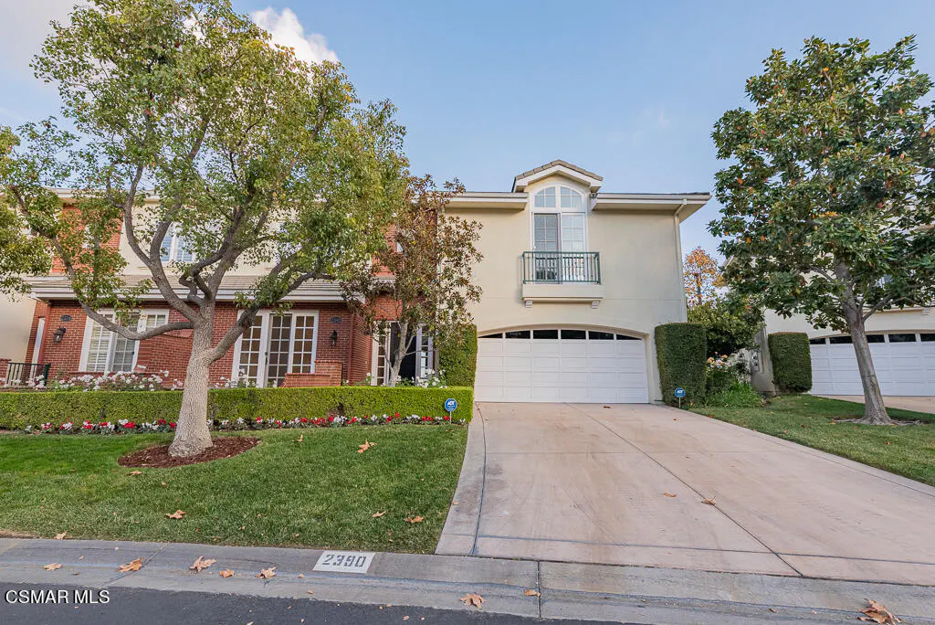 Two-story homes with white garages, brick and beige exteriors, green lawns, and trees under a blue sky.