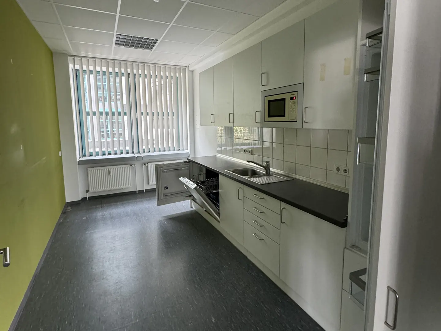 A bright kitchen with white cabinets, a microwave, and a sink. A dishwasher door is open. A window with blinds is on the left.