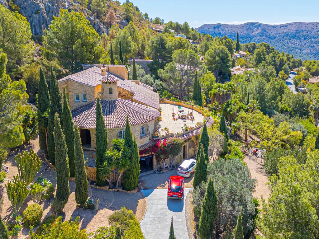 Aerial view of a stone house with a red tile roof, surrounded by trees, with two cars parked in the driveway.