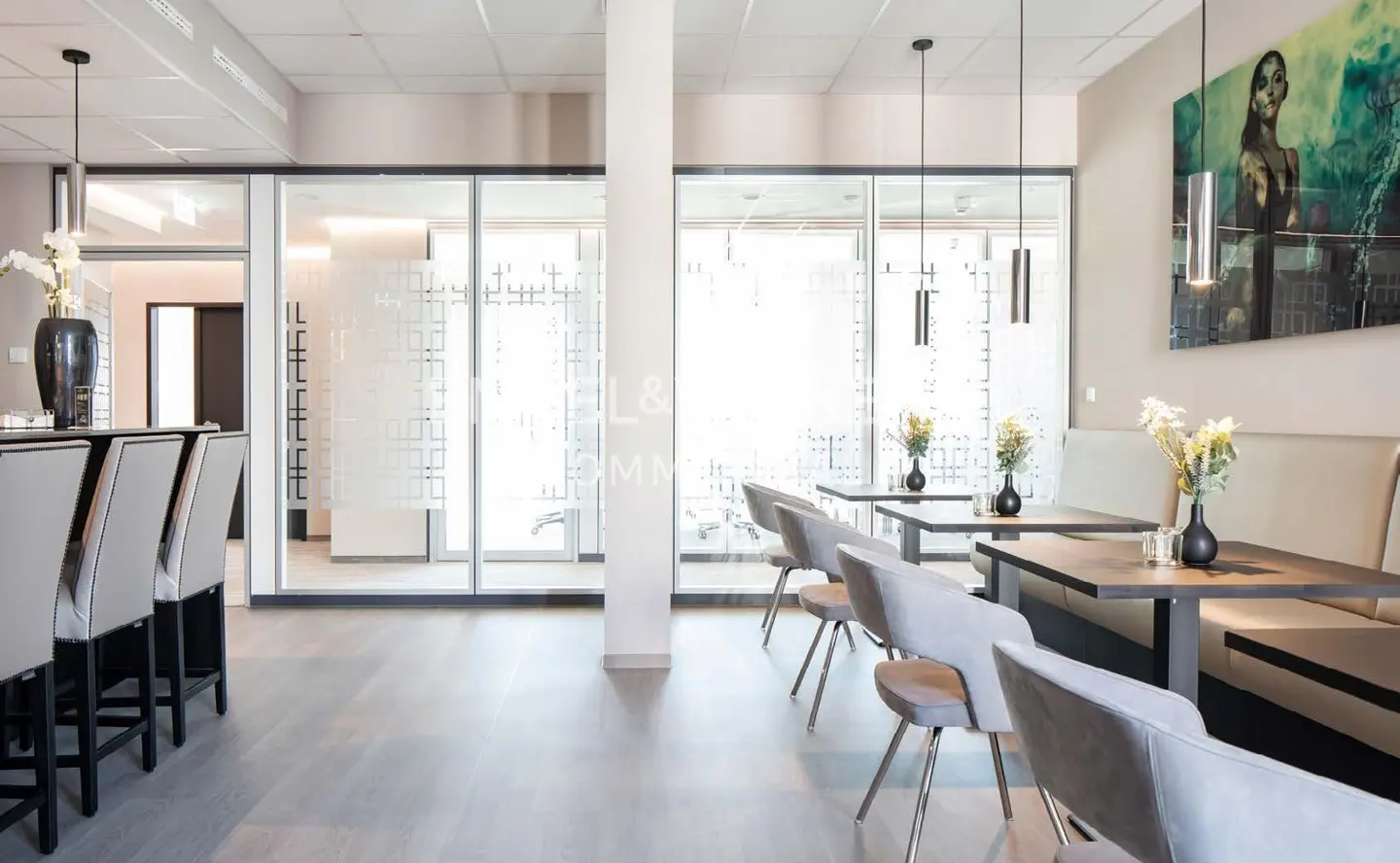 Modern office space with bar stools, tables, and chairs. Glass-walled offices are visible in the background. A large painting hangs on the wall.