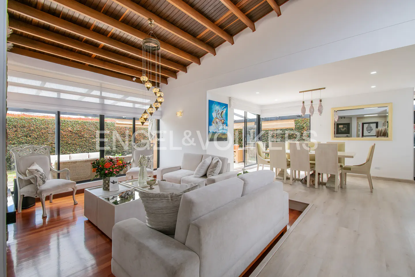 Bright, open-concept living and dining area with white sofas, wood floors, and a dining table with six chairs. Large windows overlook a green hedge.