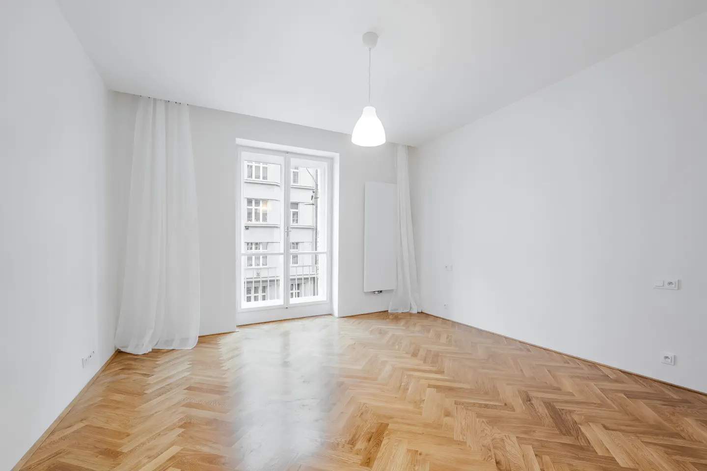 Bright, empty room with herringbone wood floors, white walls, and a window with sheer curtains. A white pendant light hangs from the ceiling.