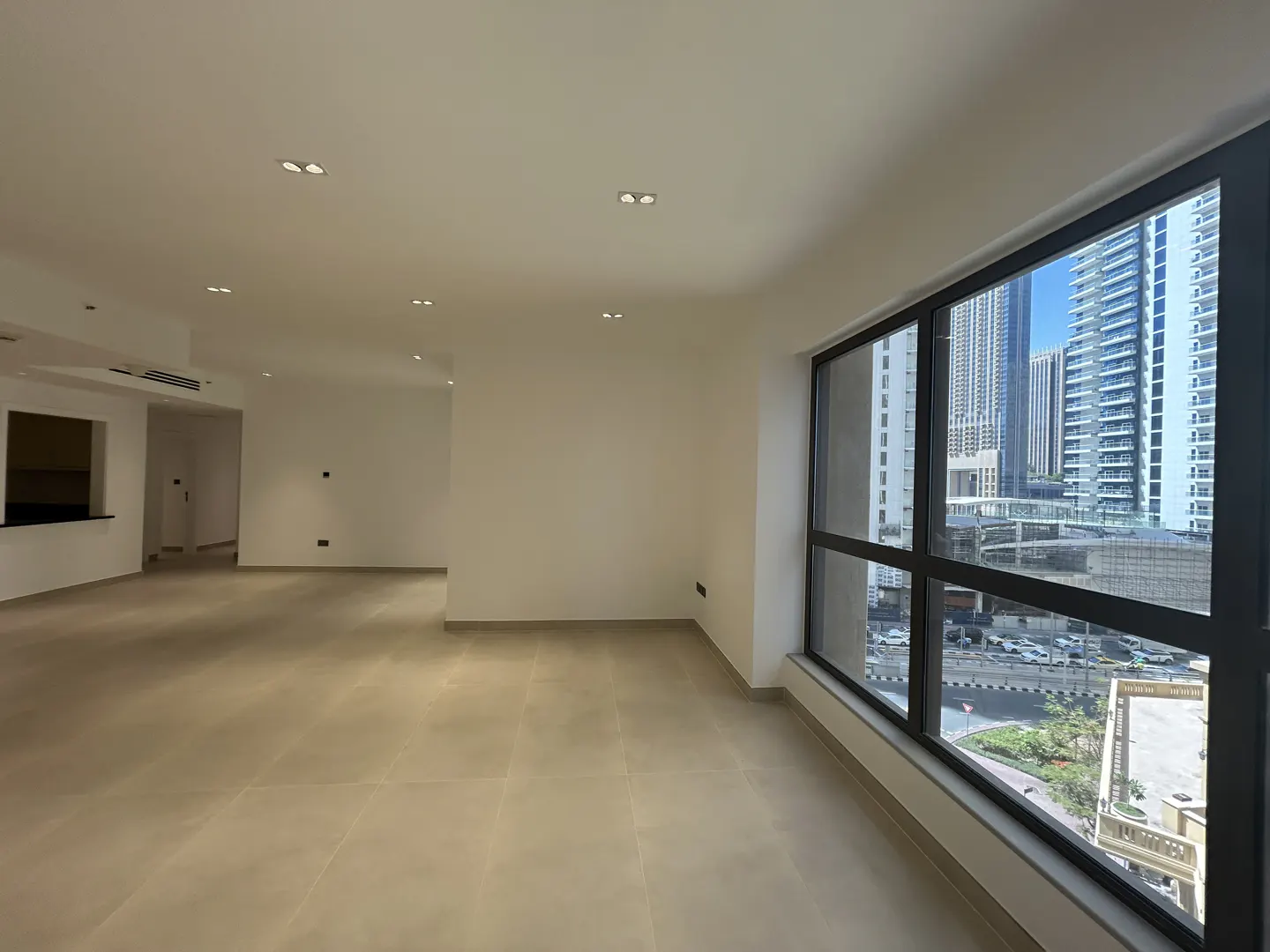 Empty, modern apartment with white walls, beige tile floors, recessed lighting, and large windows overlooking city towers.
