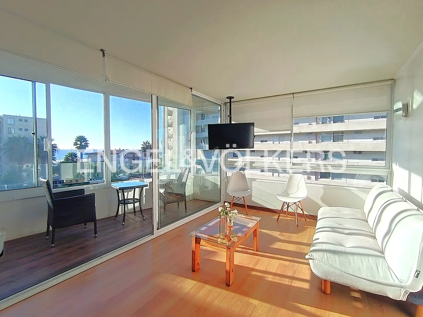 Bright living room with a white sofa, chairs, and a glass coffee table. Large windows offer ocean views. A flat-screen TV hangs from the ceiling.