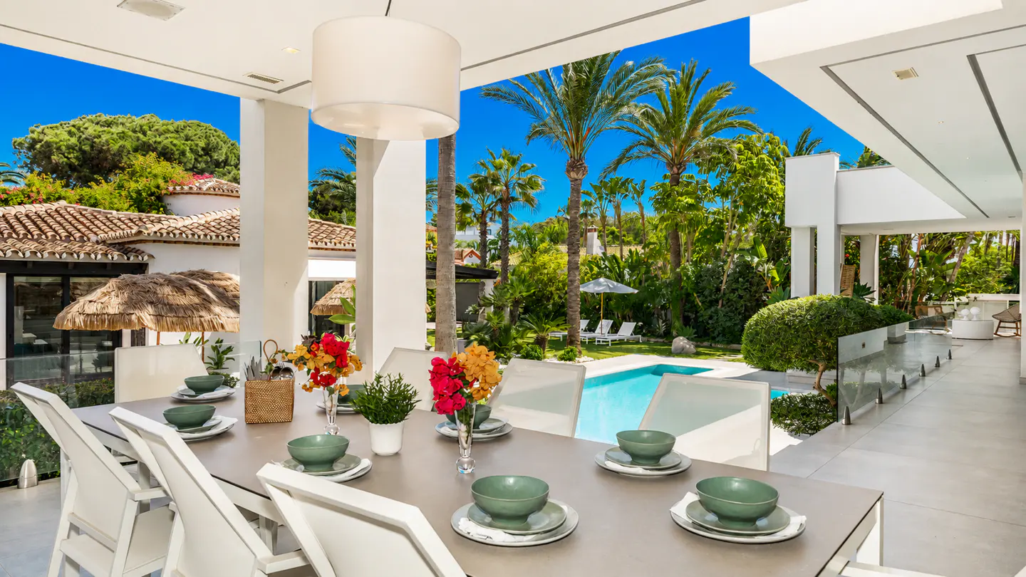 Outdoor dining area with a long table set for a meal, overlooking a pool and lush greenery.