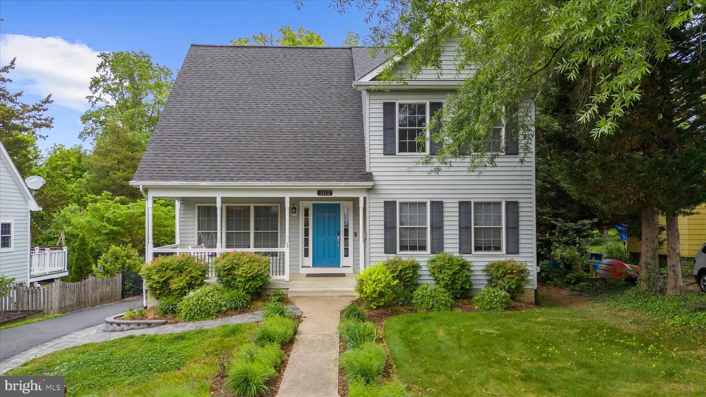 Two-story light gray house with a dark gray roof, black shutters, and a bright blue front door. A small porch is on the left. Green lawn and trees.